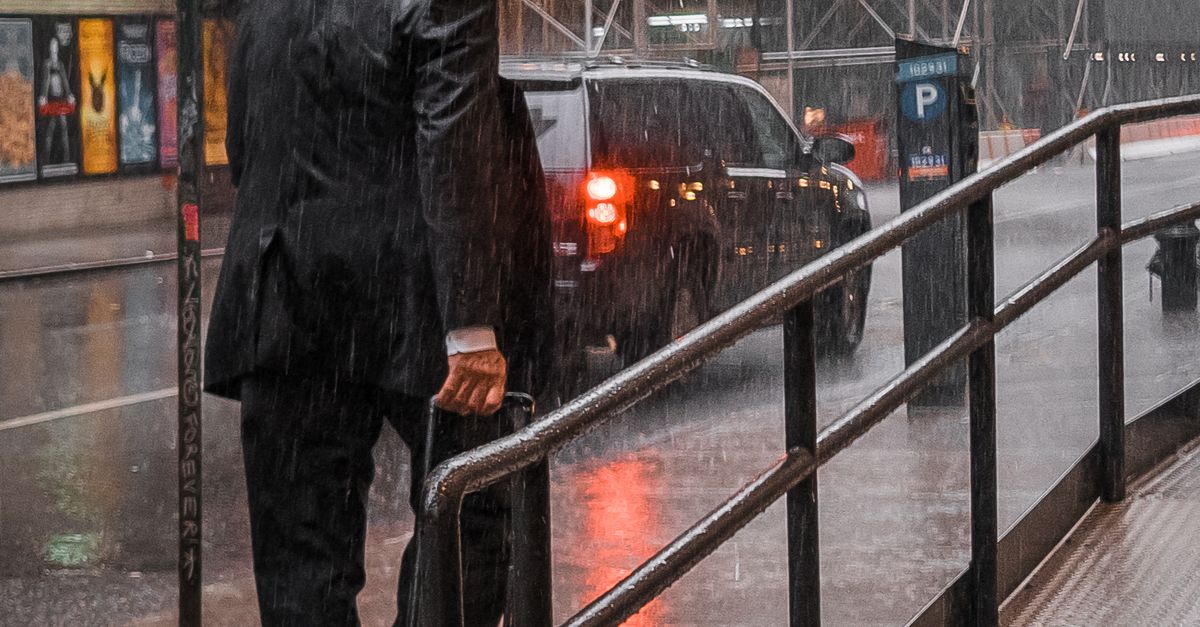 Rüyada Yağmur Altında Yürümek - Rüya Tabiri ve Anlamı - A businessman walks along a rainy city street with an umbrella and luggage.