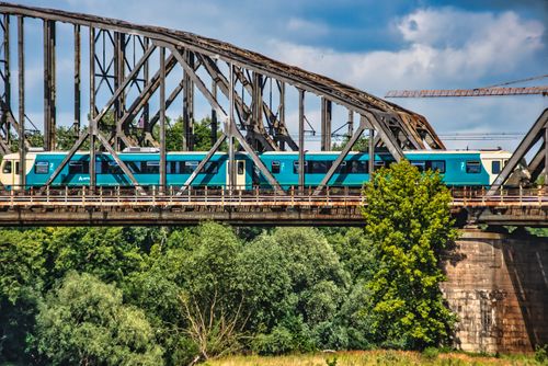 Free A turquoise train crossing an iron bridge amidst lush greenery in Toruń, Poland. Stock Photo