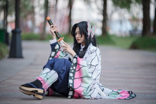 A cosplayer in a colorful butterfly-themed kimono sitting outdoors holding a sword.