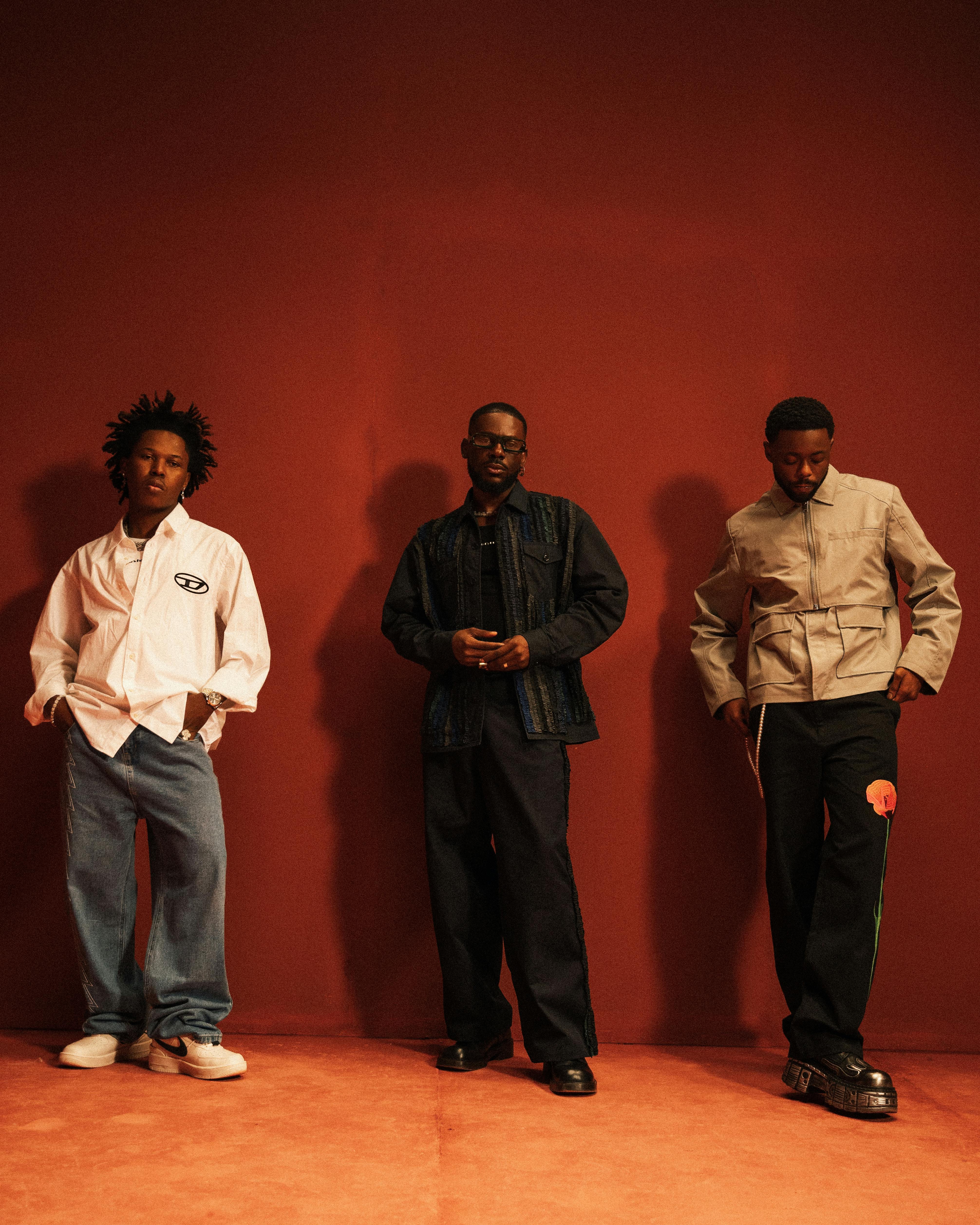 Three men pose against a warm red background in fashionable casual wear, embodying style and confidence.