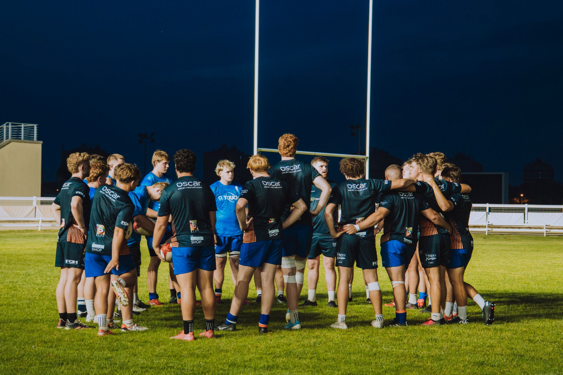 Rugby players gather in a team huddle under stadium lights at night.