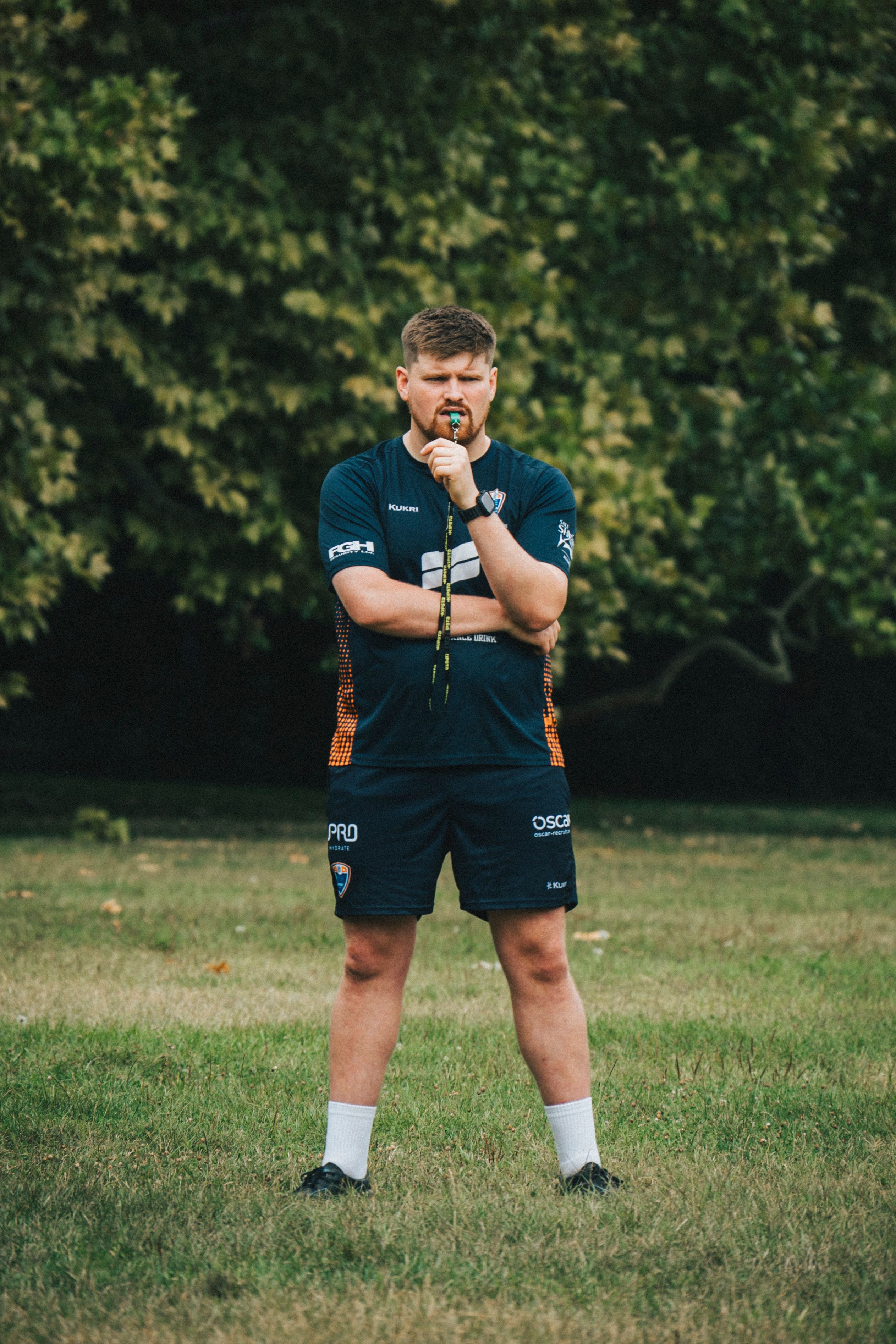 Male sports coach in athletic gear holding a whistle while standing on grass outdoors.
