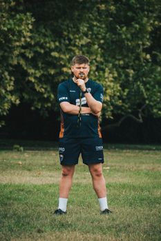 Male sports coach in athletic gear holding a whistle while standing on grass outdoors.