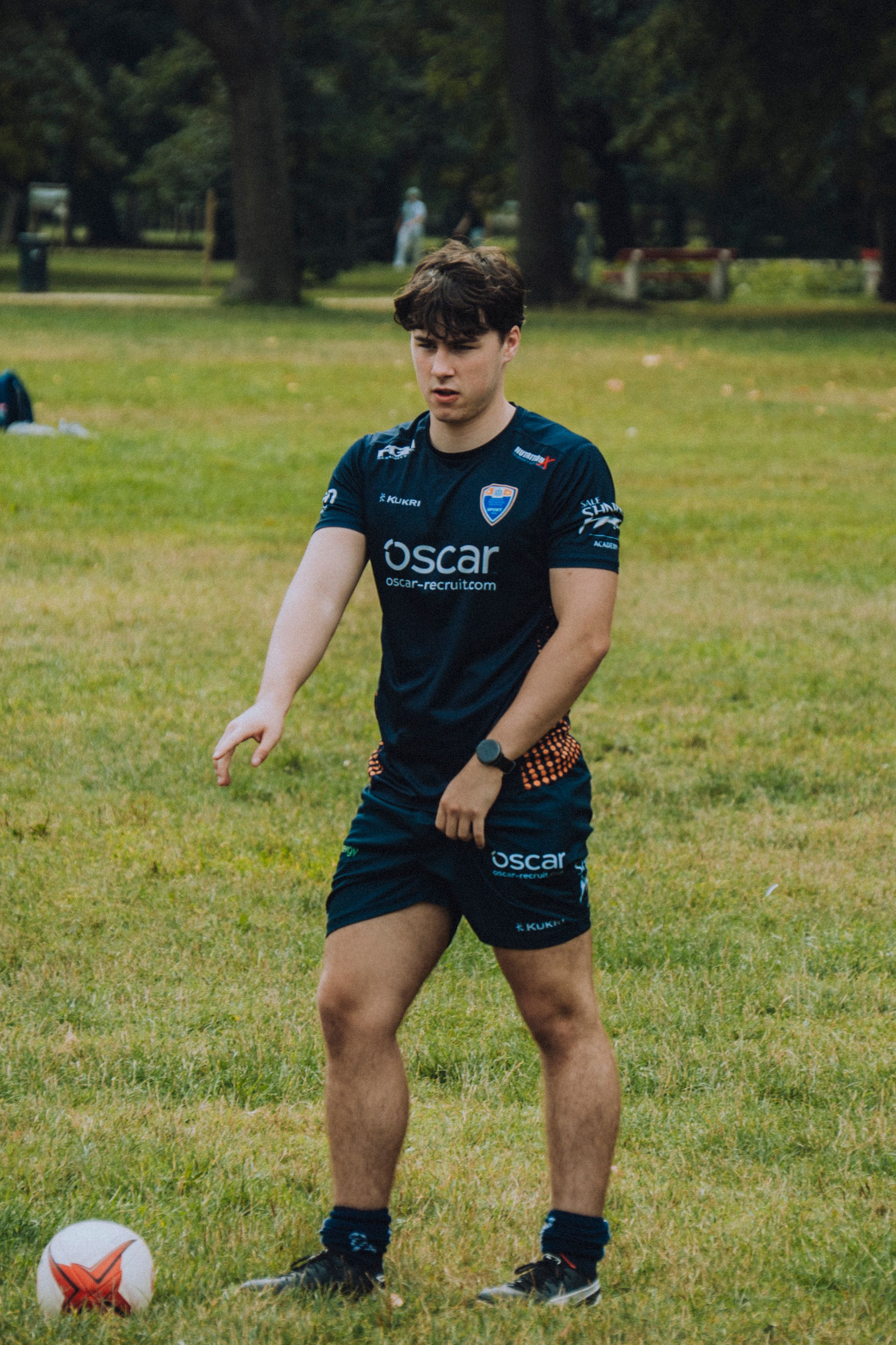 A young adult male practices rugby skills on a grassy field, wearing a sports uniform.