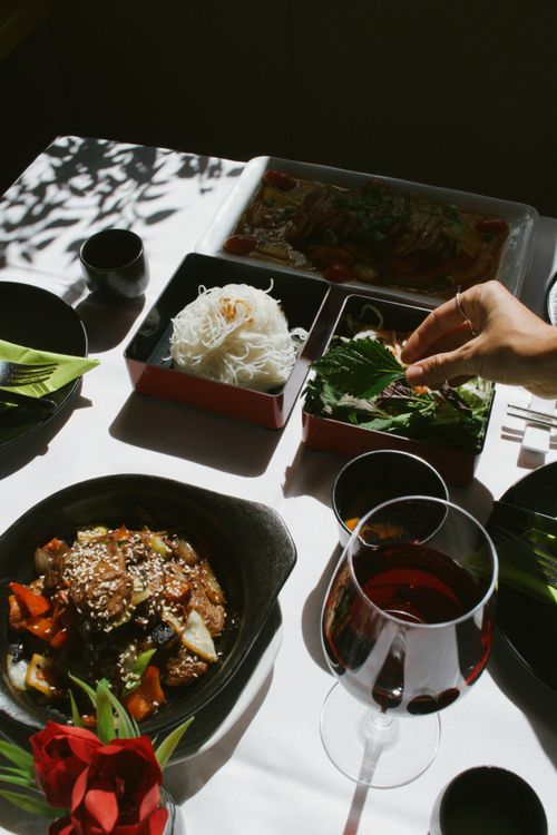 Free A sunlit table with Asian dishes, fresh greens, and a glass of red wine creates an inviting culinary ambiance. Stock Photo