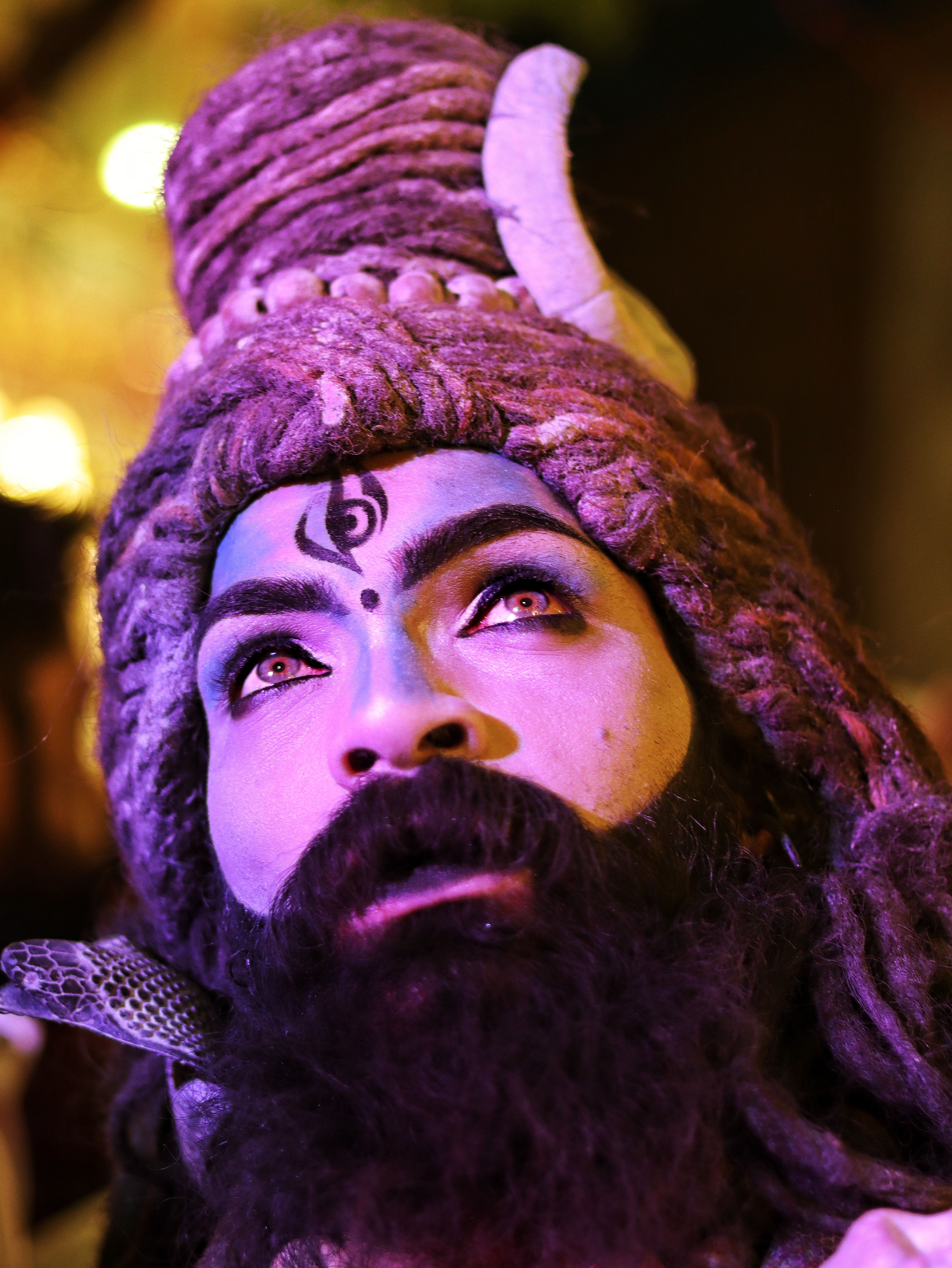 Close-up of a man impersonating Lord Shiva, with intense makeup and dreadlocks, illuminated by vibrant lighting.