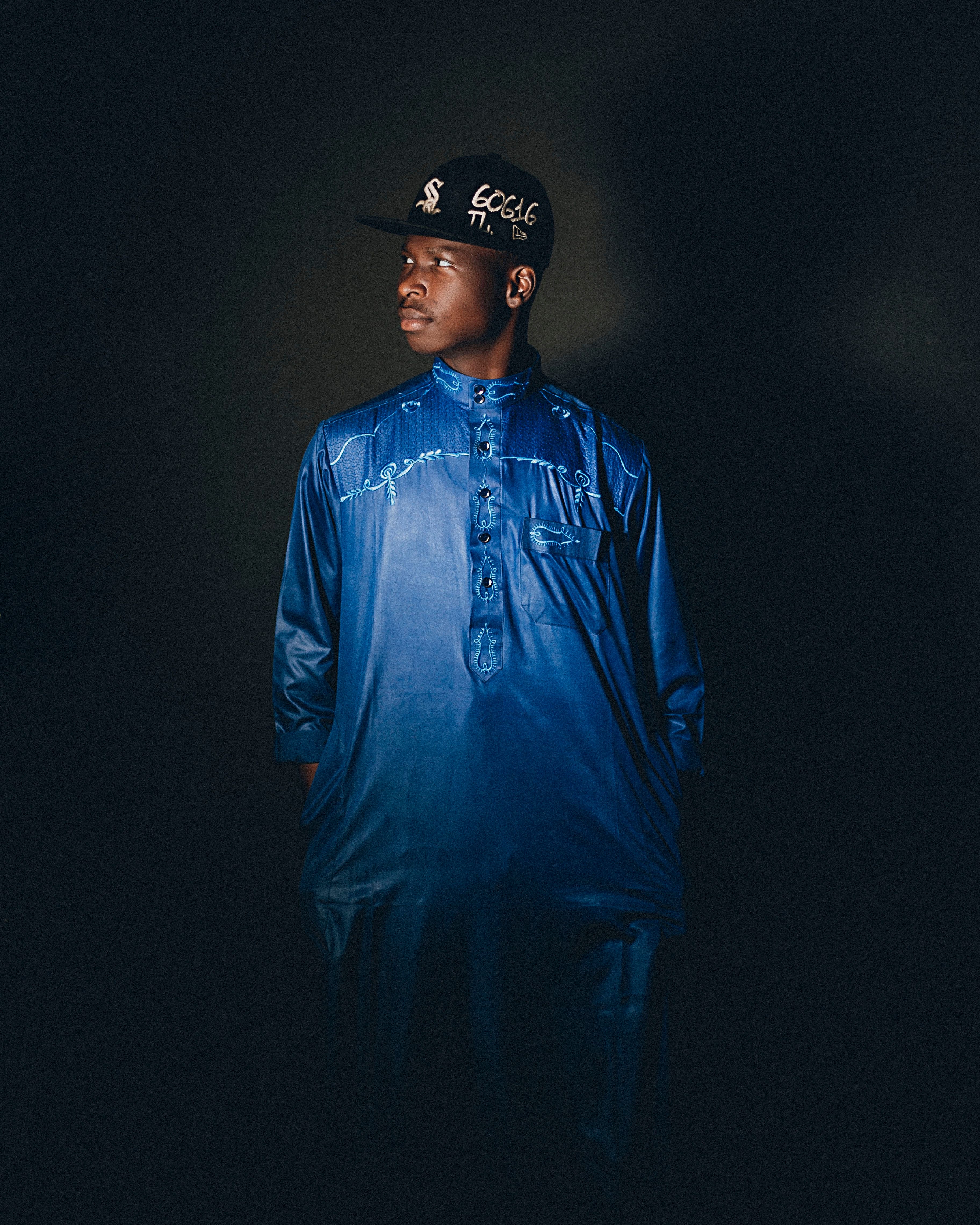 Young African man in blue traditional attire, looking to the side, dark background.