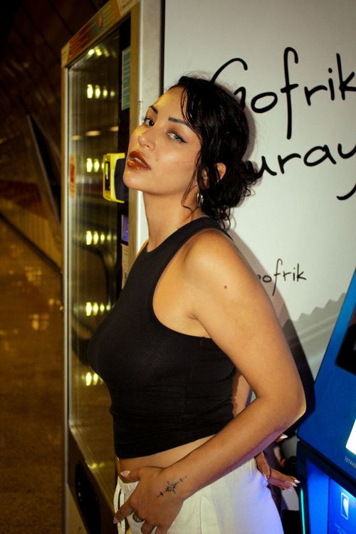 Free Woman in black top standing confidently by a vending machine with vibrant lighting. Stock Photo
