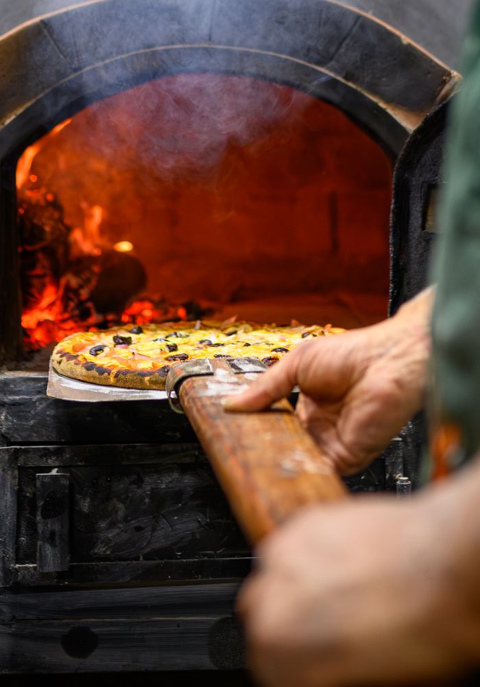 Una pizza sulla pala dentro il forno a legna, con la fiamma sullo sfondo