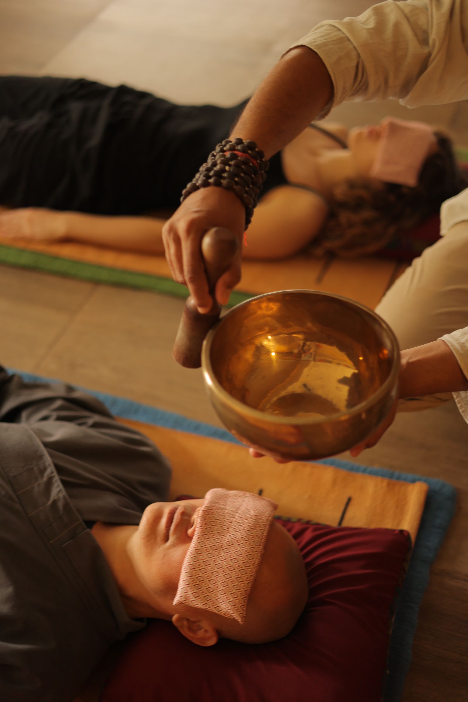 Practitioner playing a brass singing bowl beside a resting client during sound therapy