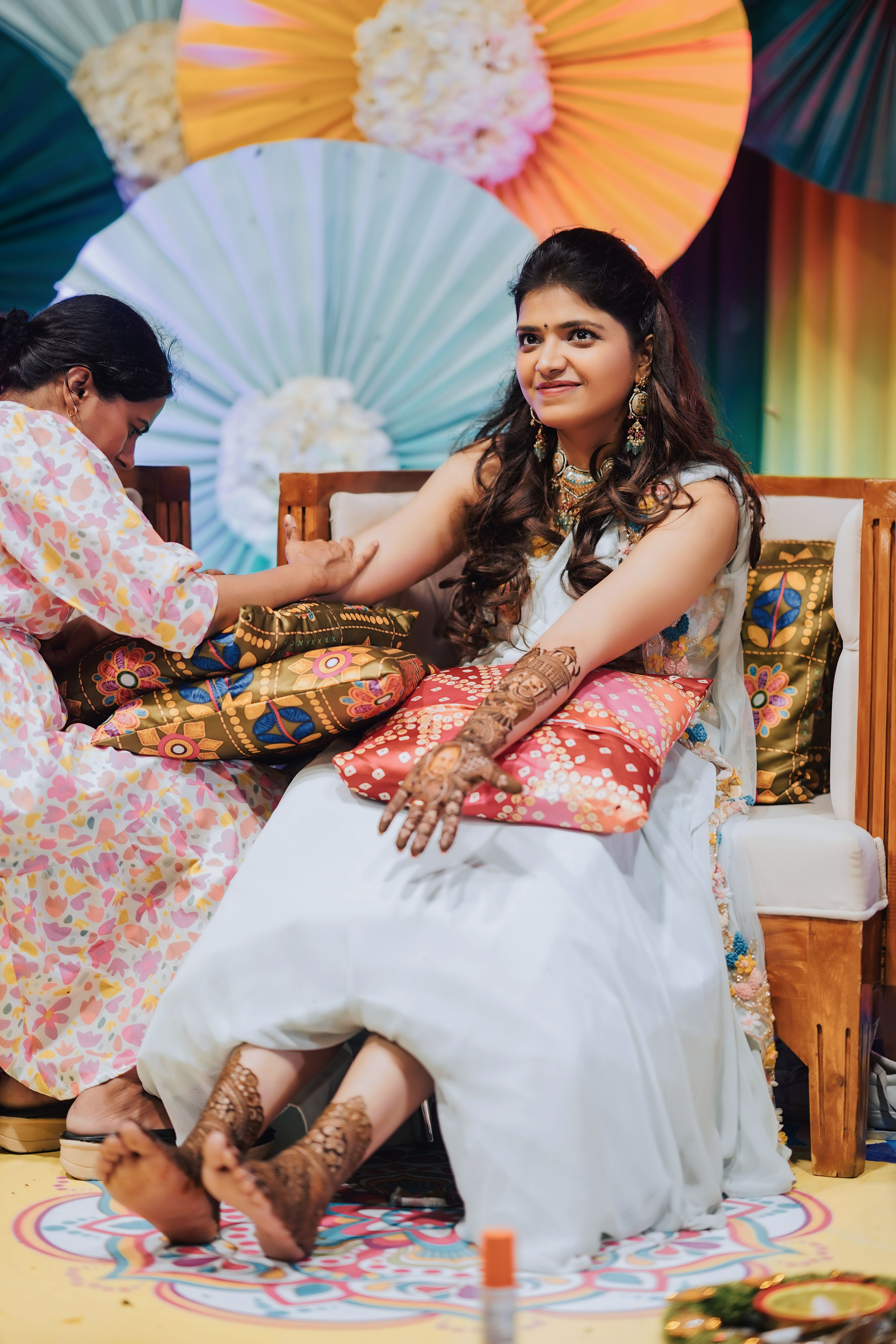 A woman gets intricate henna designs on her hands during a colorful celebration indoors.