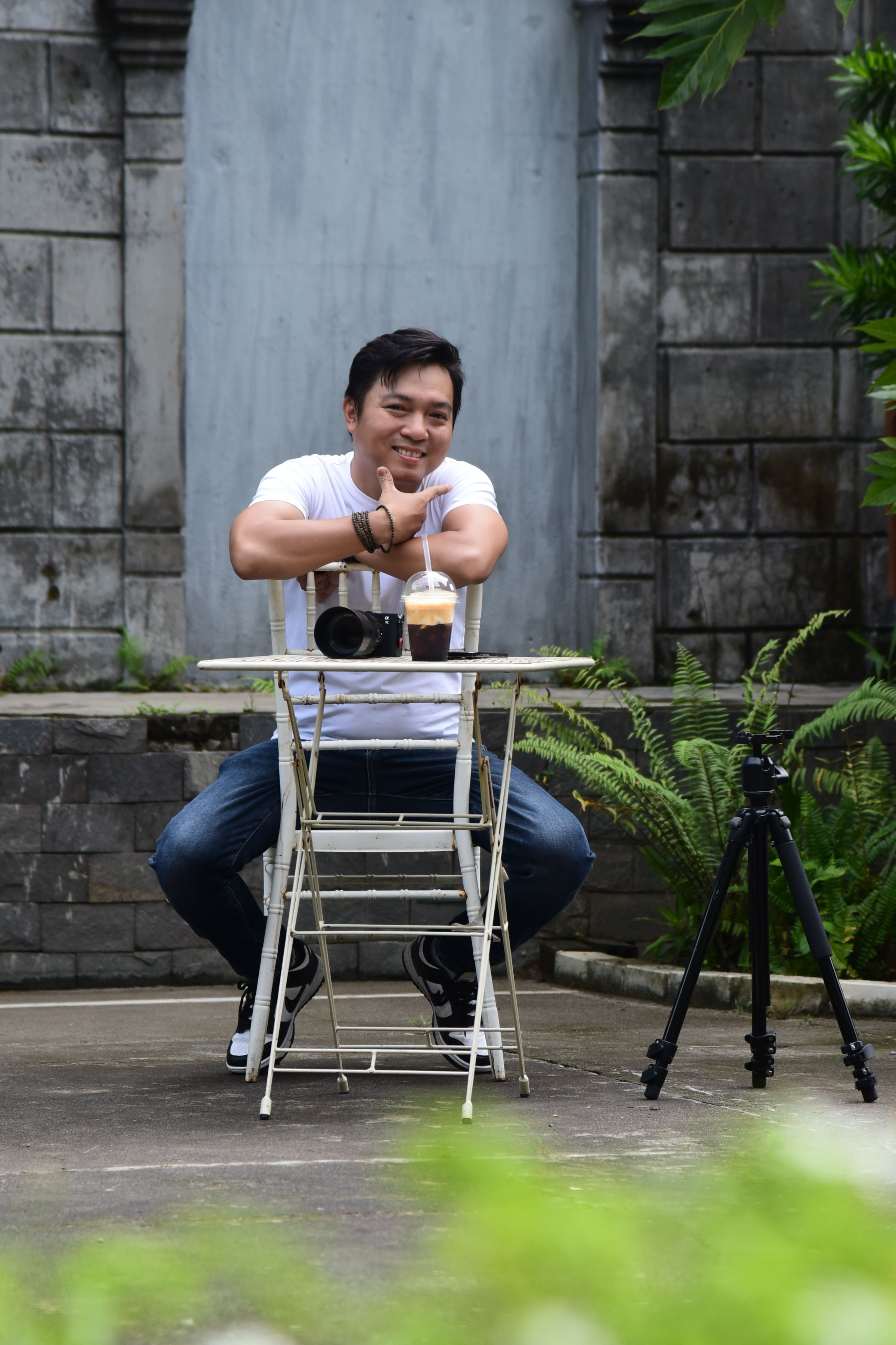 Smiling man outdoors with camera and coffee at a table. Relaxed setting.