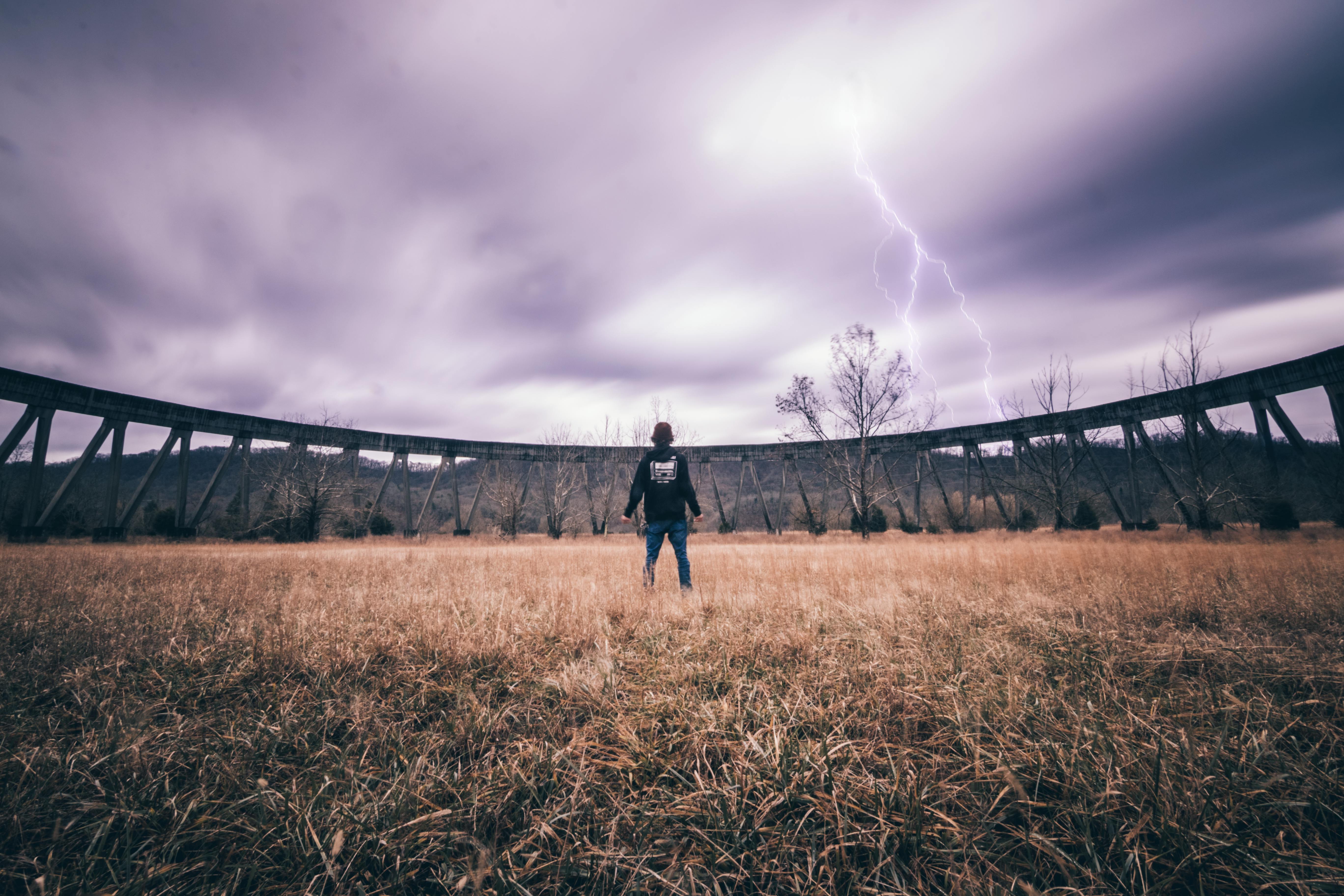A person stands in a field facing an approaching storm with visible lightning striking the sky.