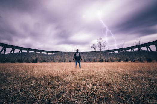A person stands in a field facing an approaching storm with visible lightning striking the sky.