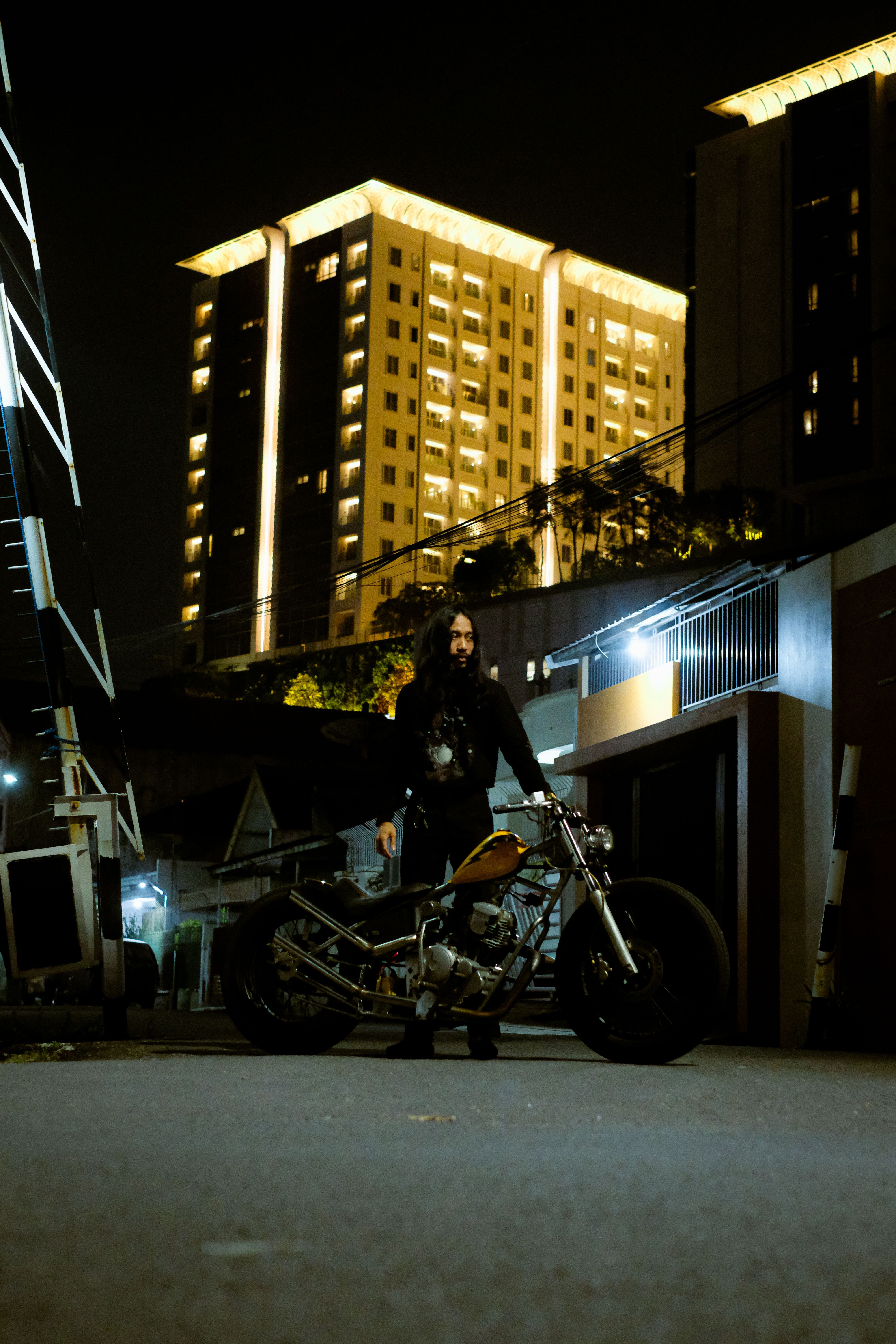 A person standing with a motorcycle in a city alley at night, with lit buildings in the background.