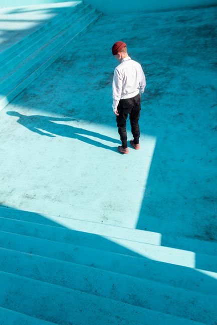 Stylish man in red beanie standing in an empty blue pool casting a shadow.