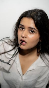 Close-up portrait of a woman with brown eyes holding sunglasses in her mouth, wearing a white shirt.