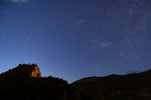 A breathtaking view of a clear starry sky with a meteor streak above a silhouetted mountain landscape at night.
