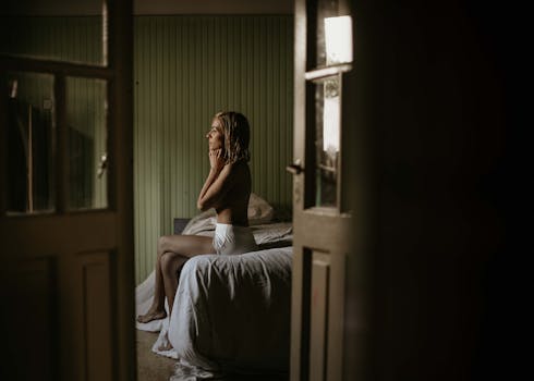 A woman sitting thoughtfully on a bed in a softly lit bedroom.