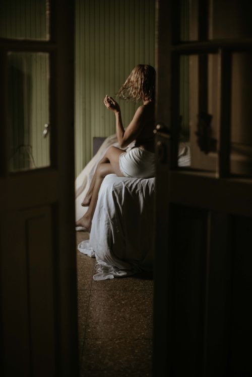 Free A woman wrapped in a sheet sits contemplatively on a bed in a dimly lit room. Stock Photo