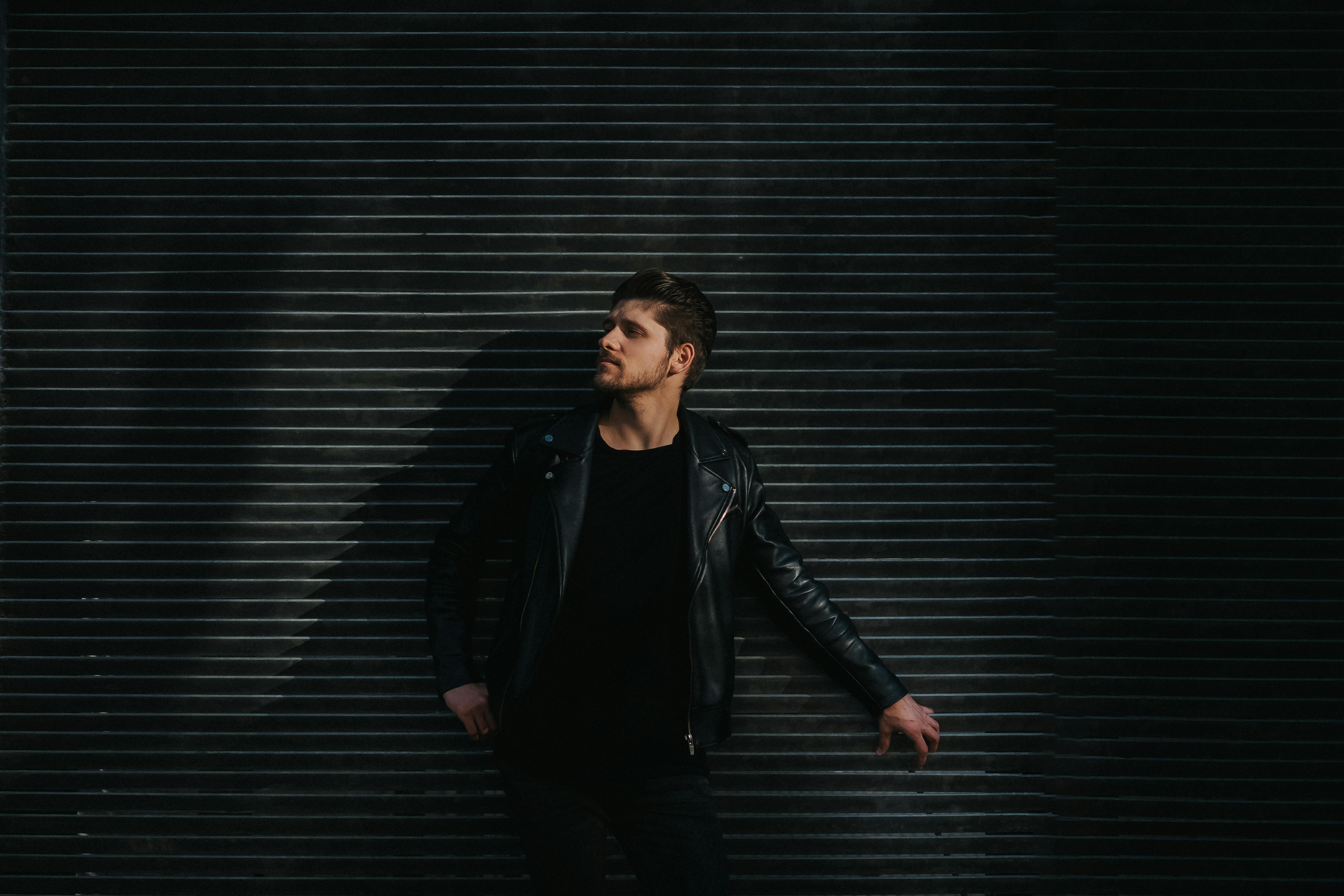 一名穿着时髦黑色皮夹克的成年男性，在情绪化的光线下倚靠着纹理墙壁