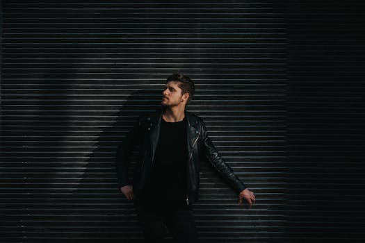一名穿着时髦黑色皮夹克的成年男性，在情绪化的光线下倚靠着纹理墙壁