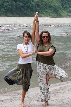 Two women practicing yoga poses by the river in Rishikesh, India, embracing wellness and nature.