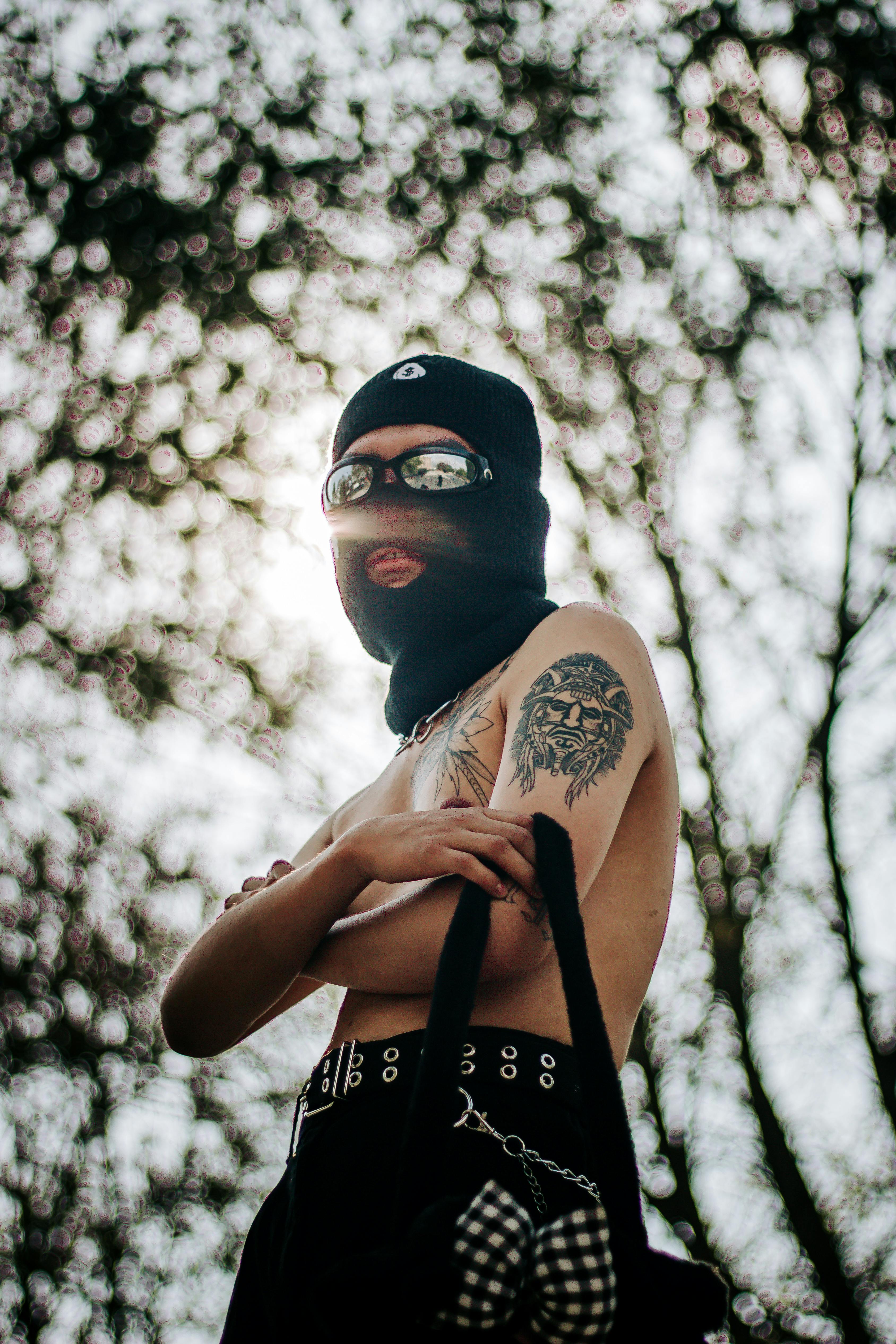 A bold portrait of a person wearing a balaclava and sunglasses, showcasing a distinctive tattoo in an outdoor setting.