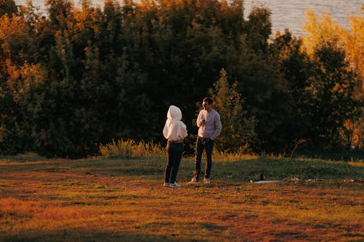 A couple stands on grassy terrain enjoying a warm autumn evening surrounded by lush trees.