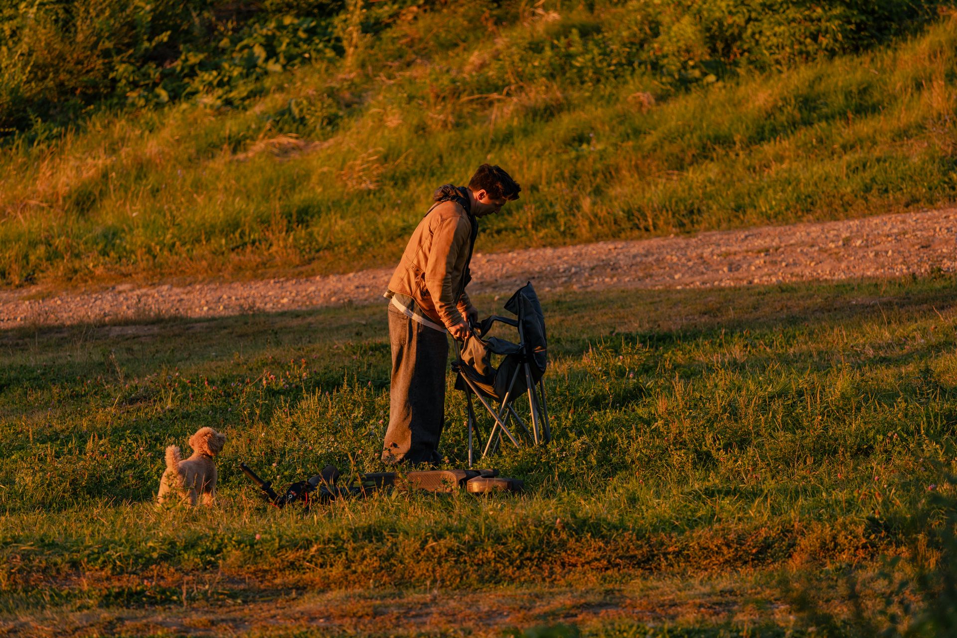 A man setting up a camping chair with a small dog nearby in a grassy field during sunset.