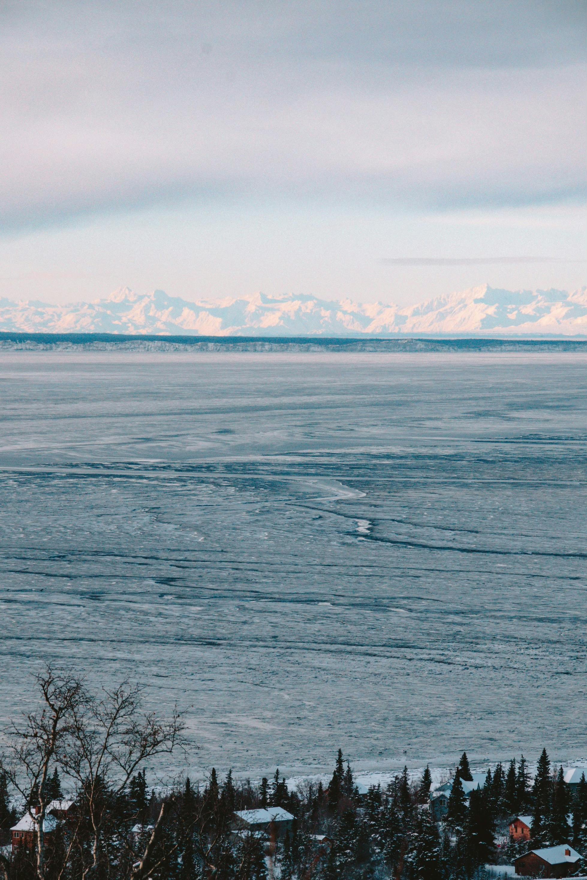 A vast frozen lake landscape with distant snowy mountains and scattered trees.