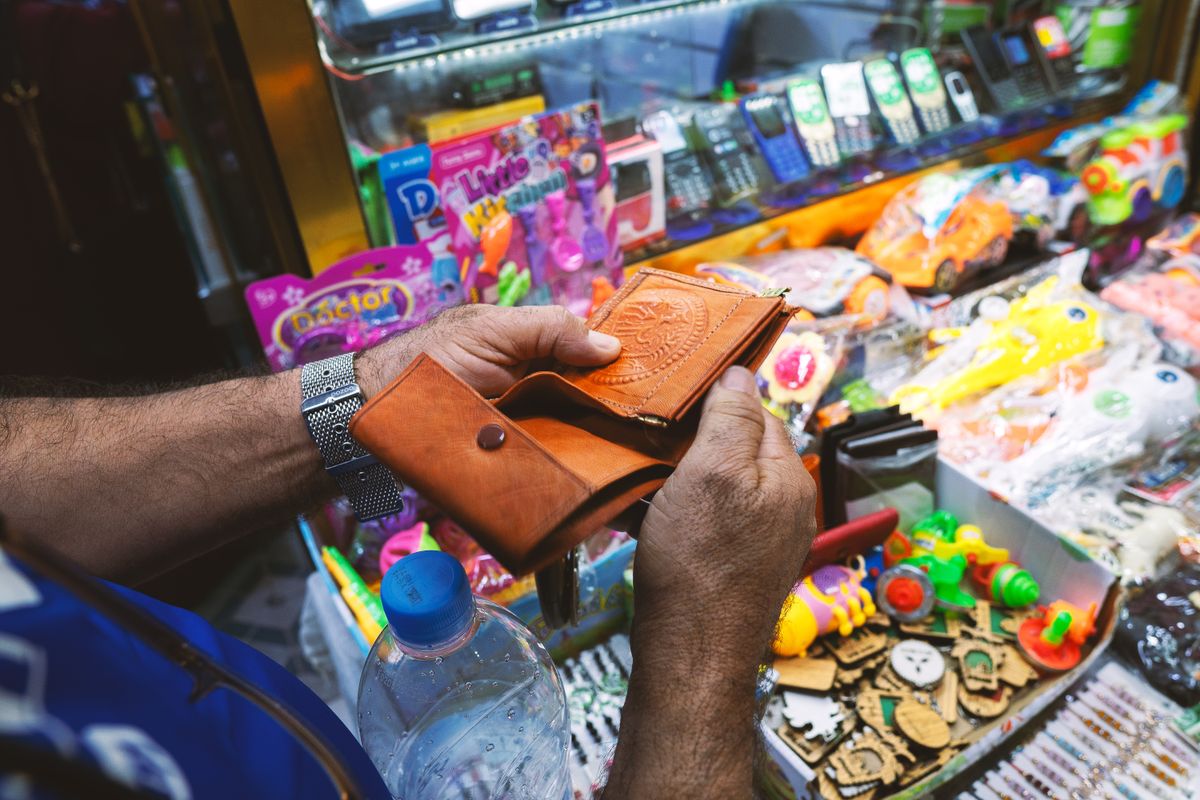 A person holding a leather wallet near a colorful toy stall in a vibrant market.