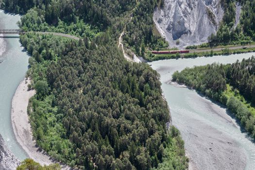 Scenic aerial view of the Rhine Gorge, Switzerland, showcasing lush forests, rivers, and a bright red train winding through the mountains.