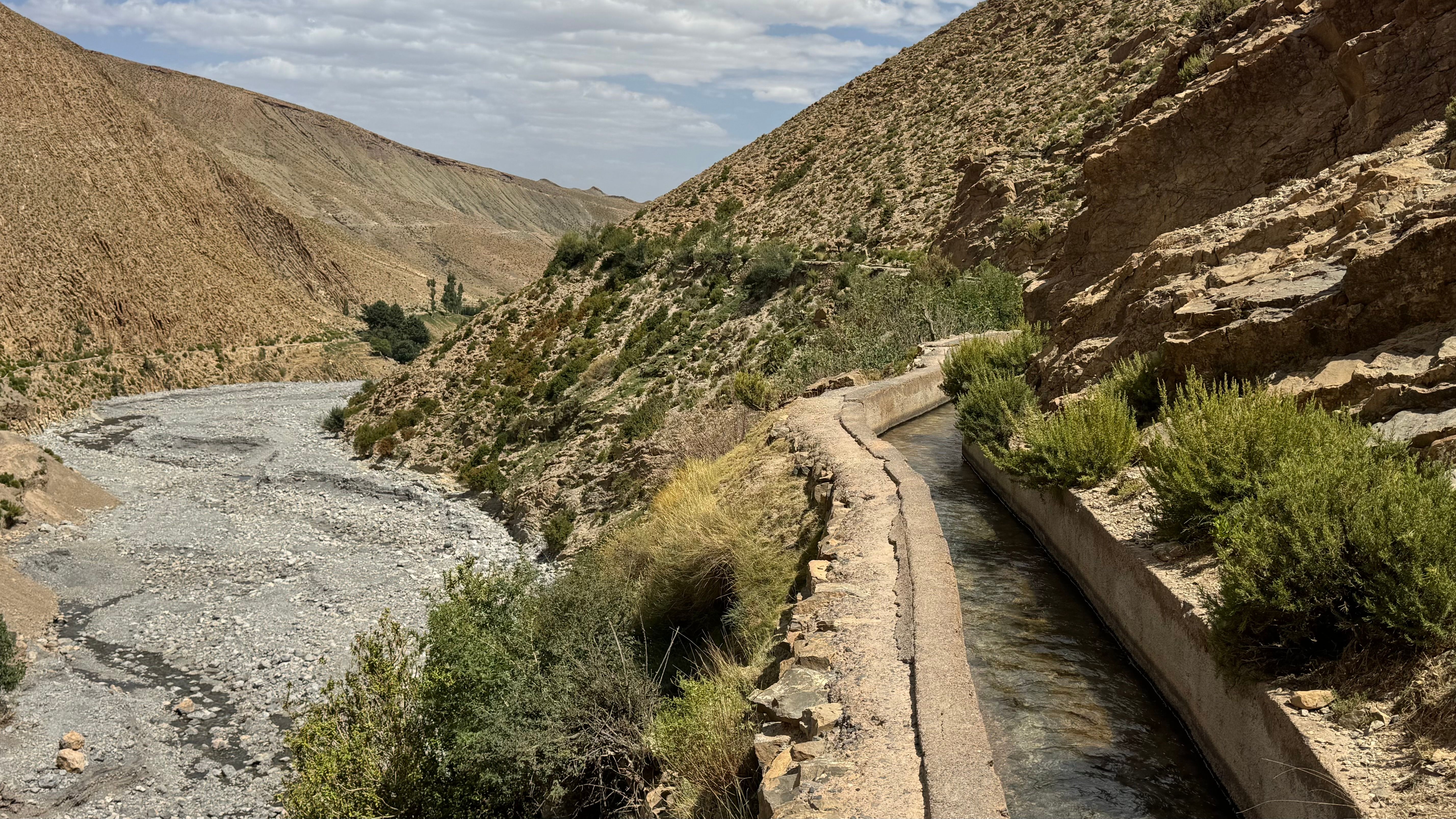 A picturesque irrigation canal winds through a dry valley in Morocco, surrounded by rugged hills under a cloudy sky. - Midelt