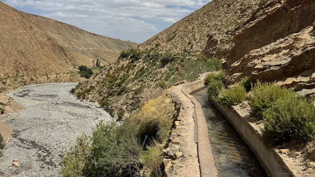 A picturesque irrigation canal winds through a dry valley in Morocco, surrounded by rugged hills under a cloudy sky.