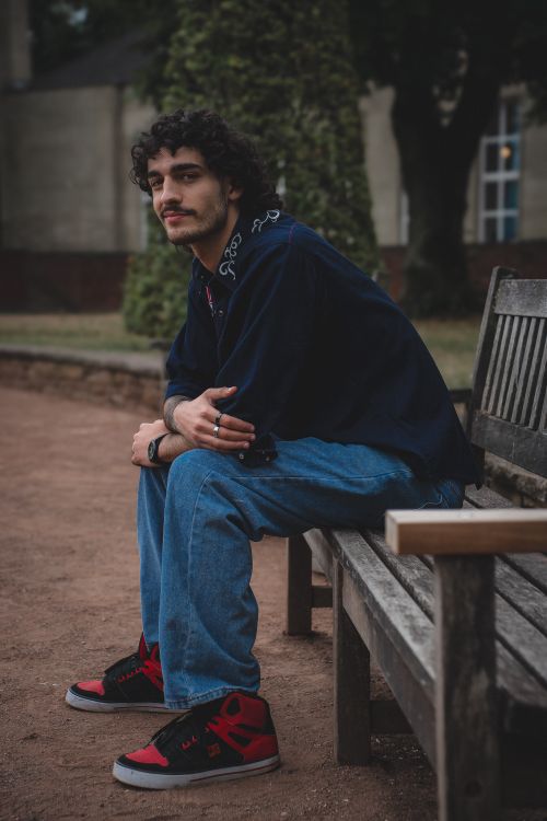 Free Casual young man with curly hair sitting on a bench in a park setting. Stock Photo