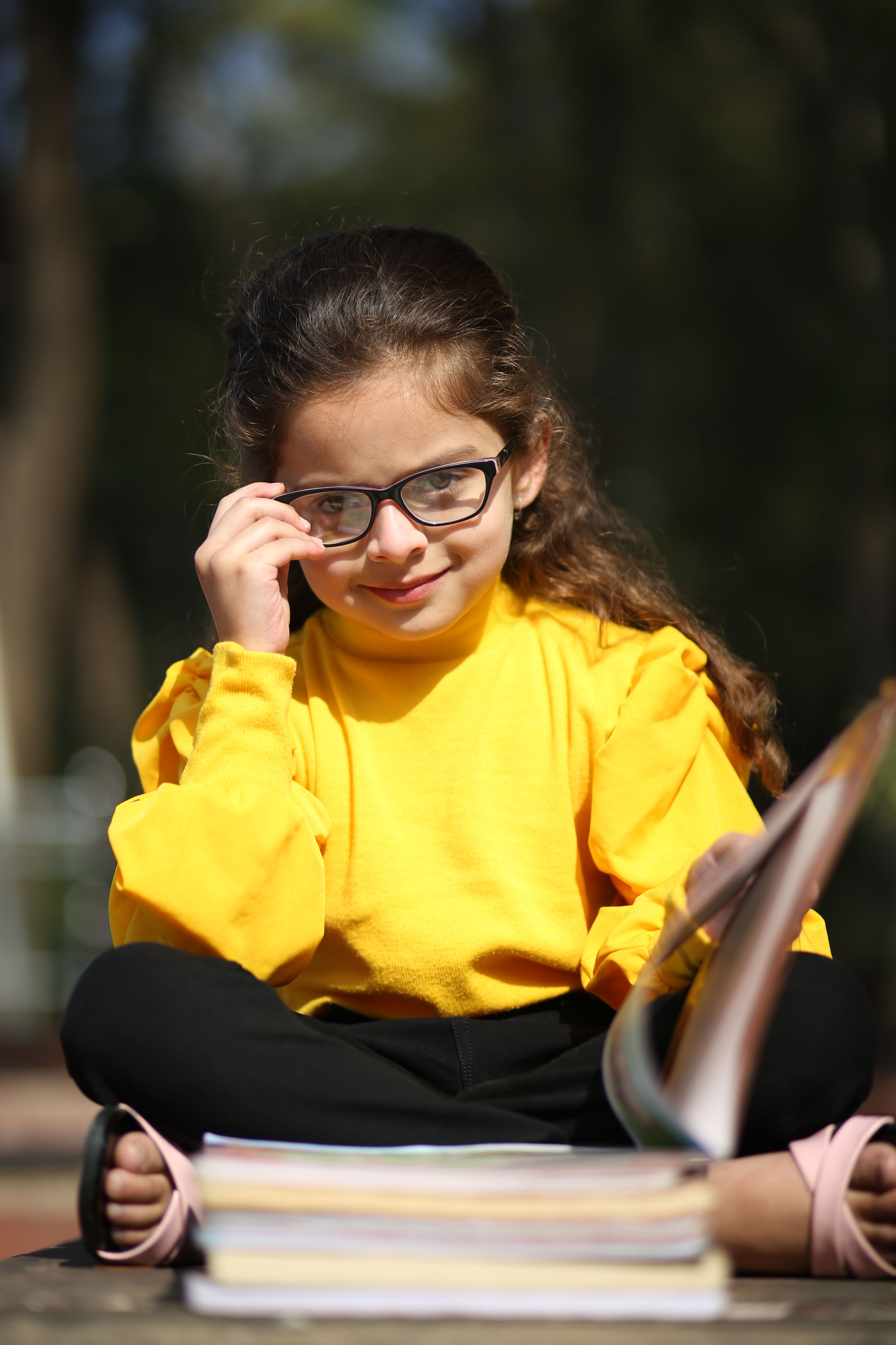 A cheerful young girl in glasses reading outdoors on a sunny day in Dewas, India.