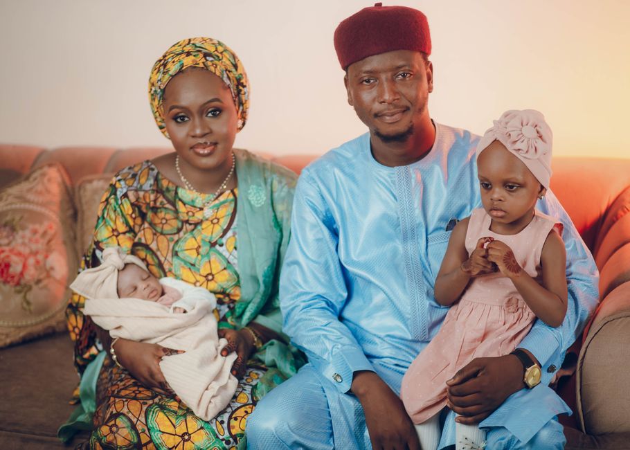 Dark-skinned Nigerian family of four gathered around a dining table in a modest Lagos home kitchen, sharing a large pot of jollof rice and fried plantain, natural window light streaming in, wooden furniture, cheerful expressions, photorealistic