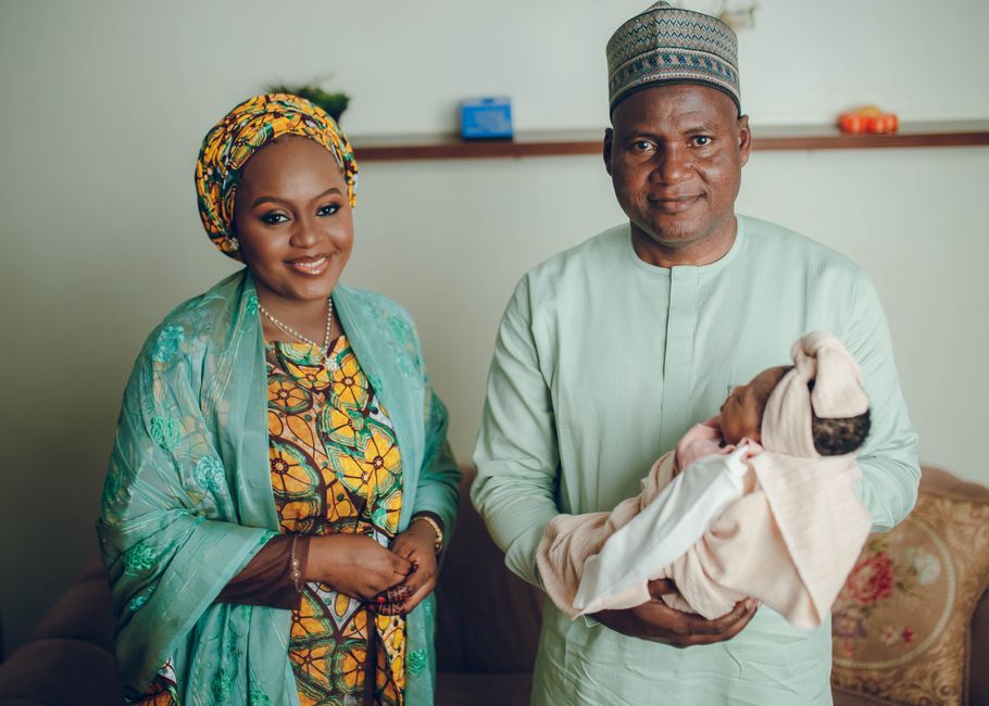 Black Nigerian family — a dark-skinned mother in colourful Ankara blouse and a father in a plain office shirt — unpacking large bags of rice, beans, and cooking oil in a modest Nigerian kitchen in Ibadan, warm indoor lighting, photorealistic, joyful expressions