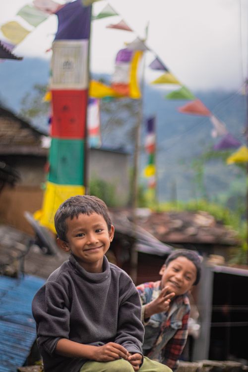 Free Two smiling children sit near colorful prayer flags in a rural mountainous area. Stock Photo