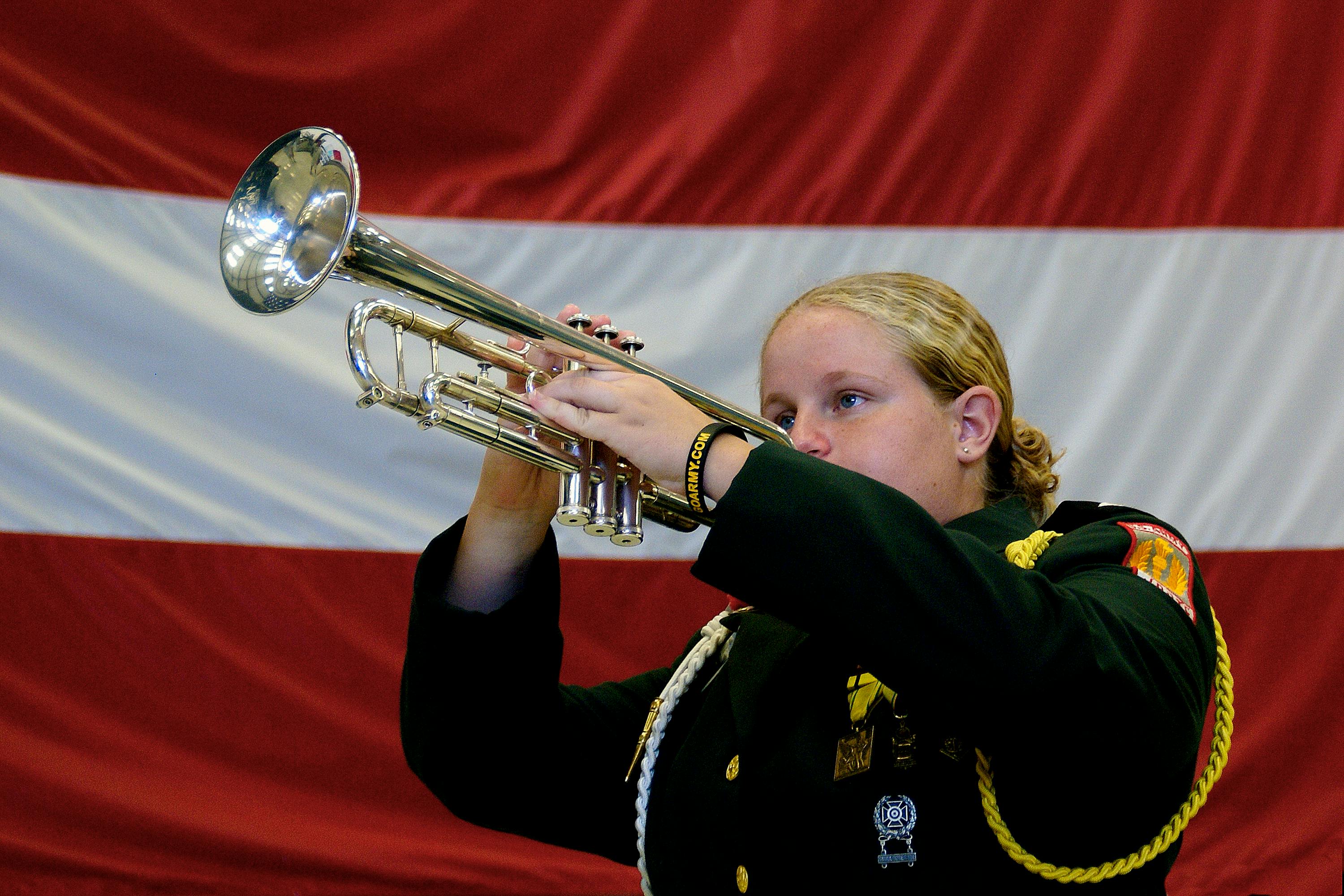 A woman in military attire plays trumpet passionately against a flag backdrop.