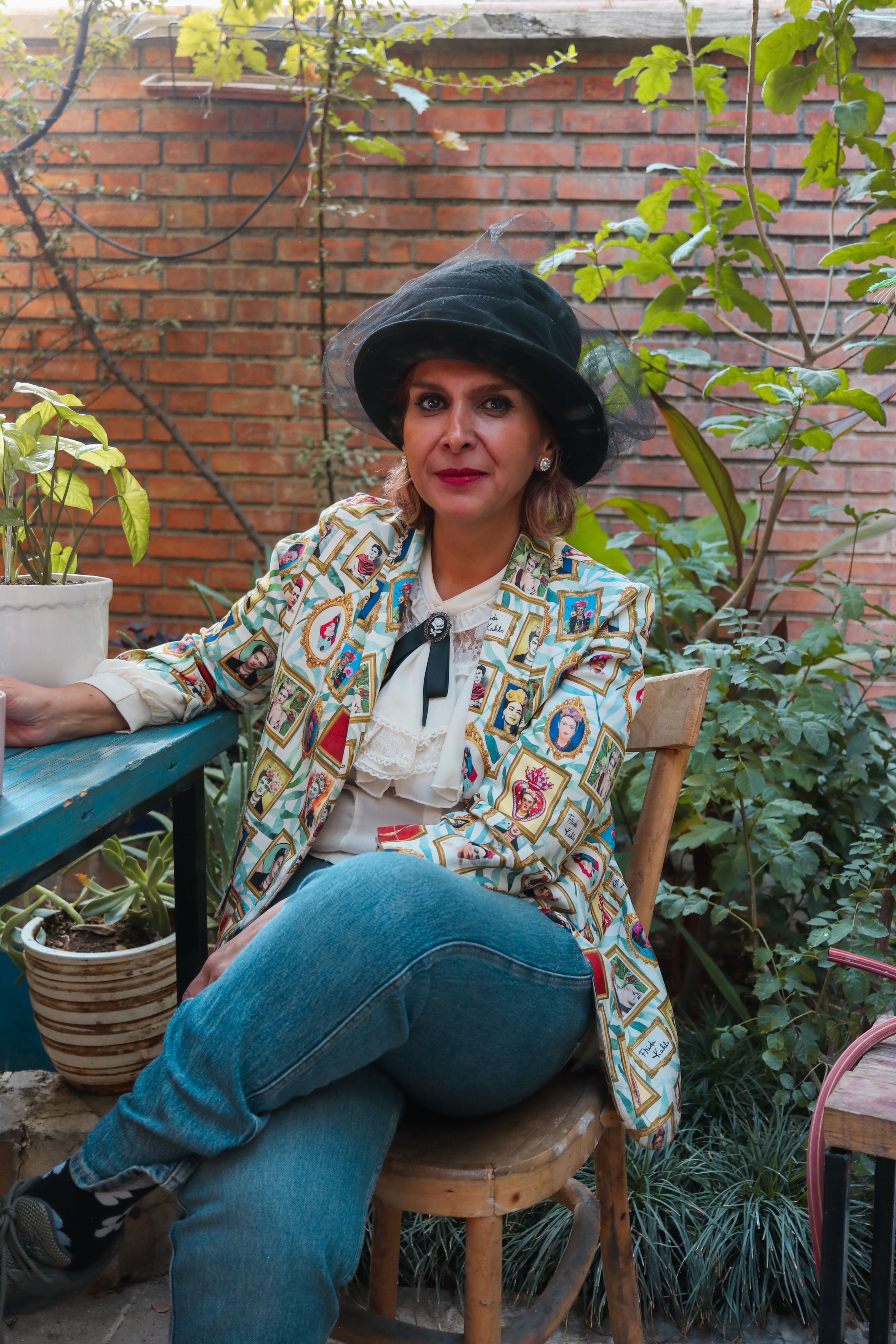 A fashionable woman sitting outdoors, surrounded by plants and brick walls.