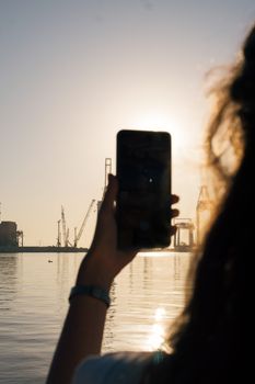 Silhouette of person with smartphone photographing a sunset at Mersin harbor with industrial cranes in the background.