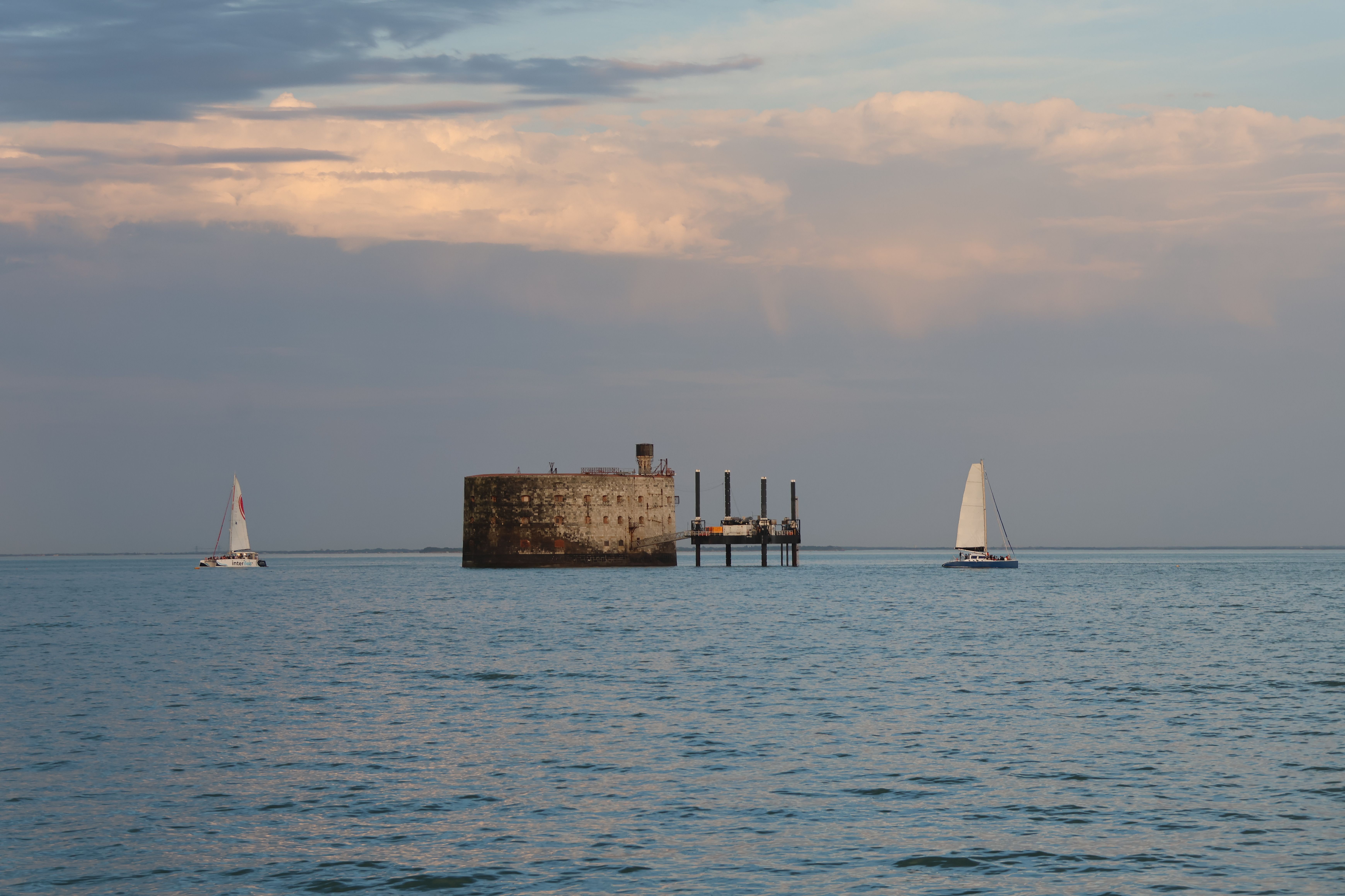Serene view of sailboats passing by a historic fort on a calm sea under a pastel cloudy sky