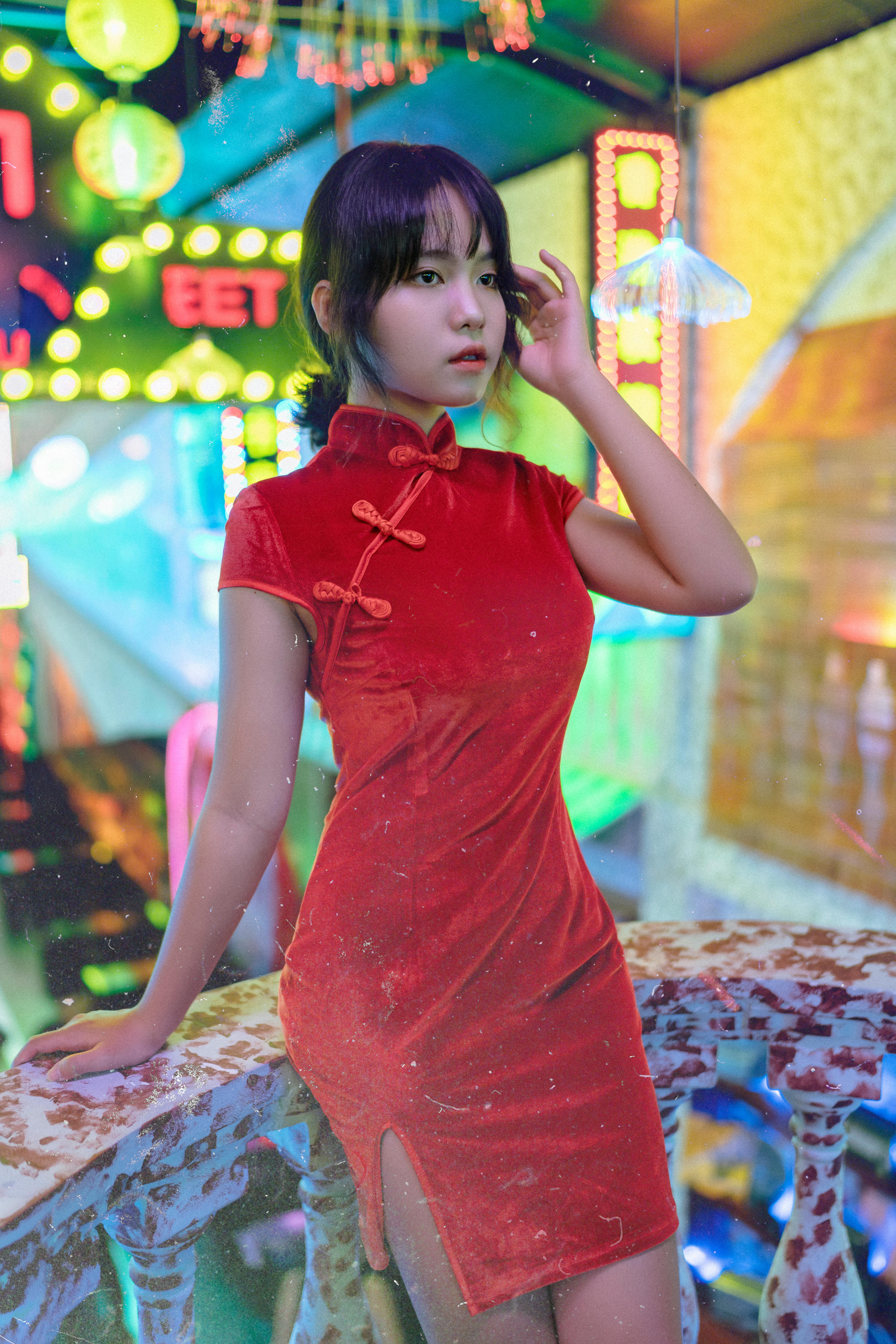 A young woman in a traditional red qipao dress poses elegantly in a neon-lit urban environment.