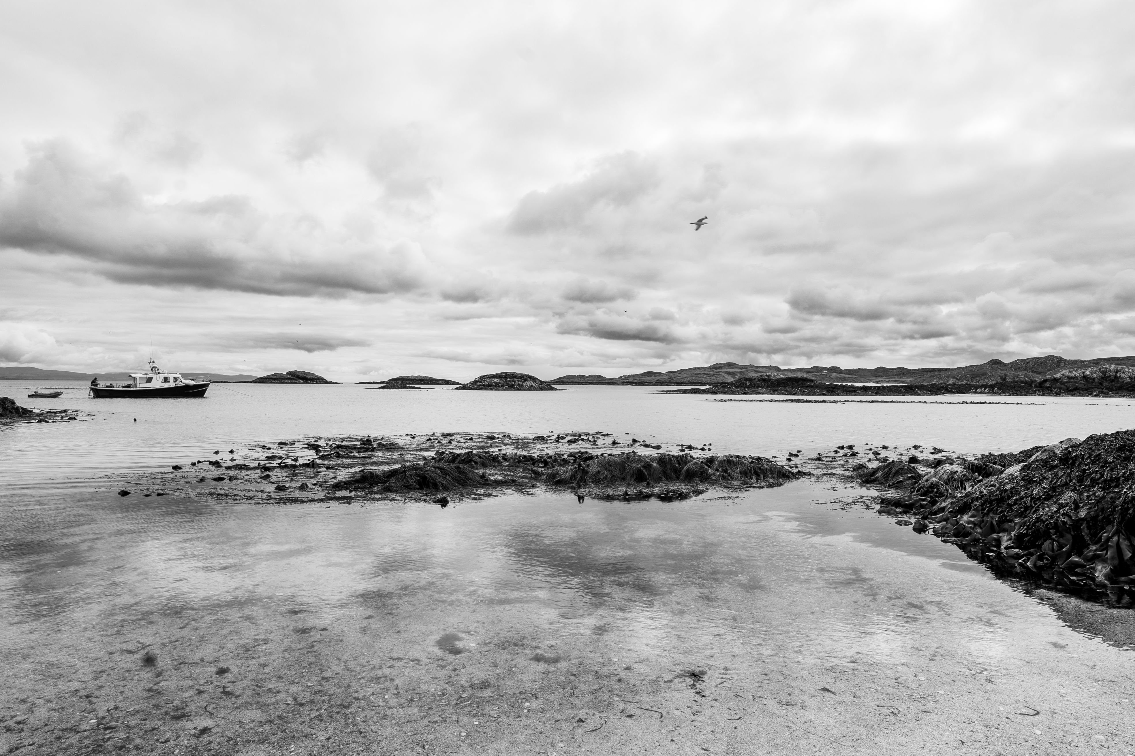 Black and white view of a calm Scottish coastline with a lone boat and dramatic clouds.