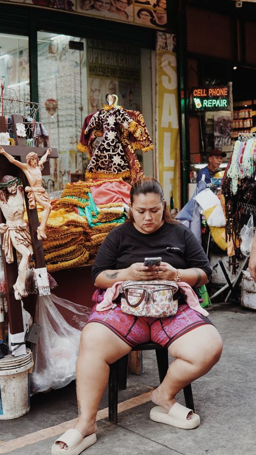 Free A street vendor sits on a chair, using a smartphone in a bustling market setting. Vibrant clothing and sale signs visible. Stock Photo