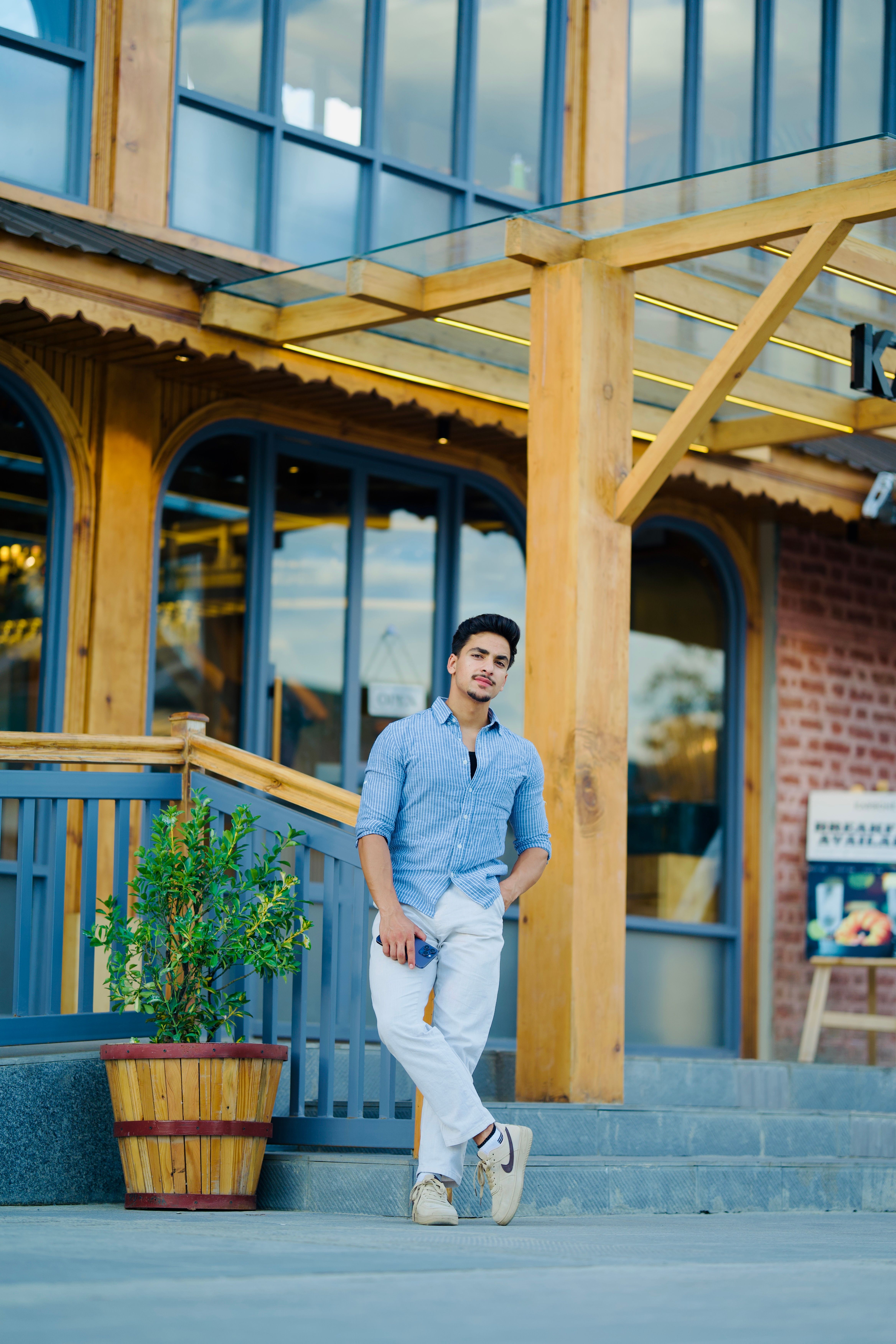 A fashionable man in a blue shirt and white pants posing confidently outside a modern café.