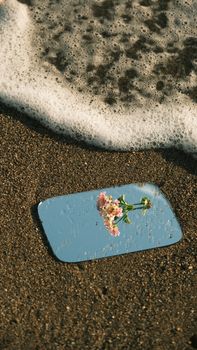 A mirror reflects a pink flower on a sandy beach near the water's edge.