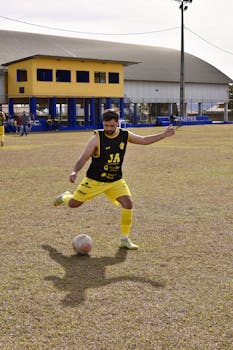 Adult male soccer player in yellow kit prepares to kick a ball on an outdoor field during daytime.